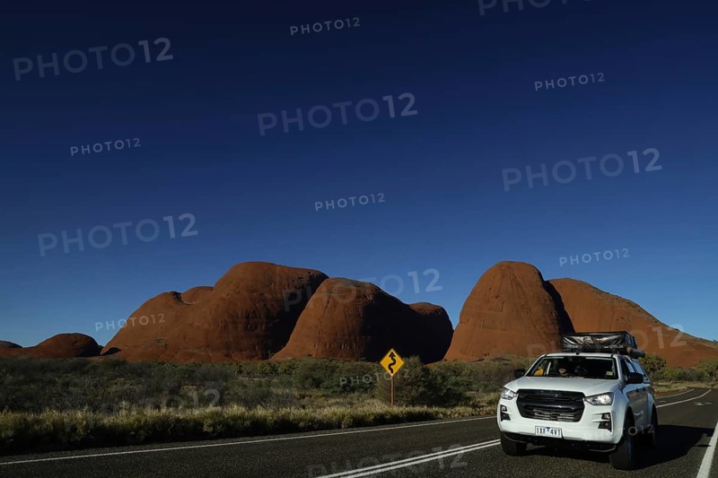 A car drives against the impressive backdrop of the Olgas under clear skies