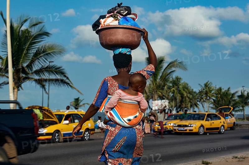 Woman carrying child on her back as palm trees and yellow taxis line the road, Abidjan, Ivory Coast