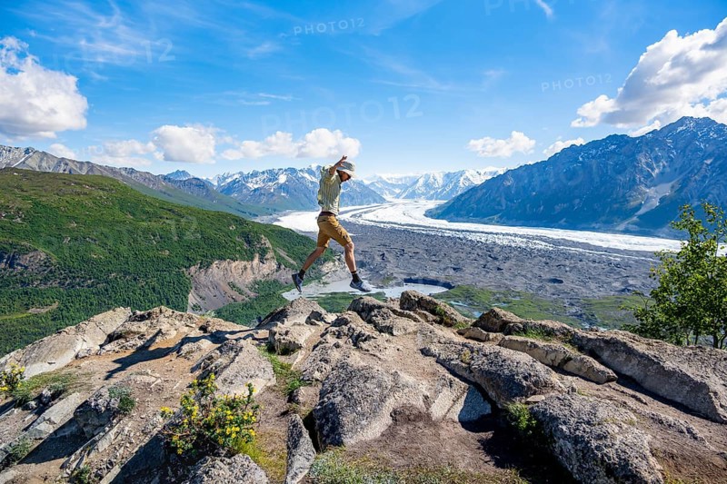 Young man jumping into the air, view of impressive mountain landscape with Matanuska glacier and glaciated mountain peaks, Lion's Head, Chugach Mountains, Alaska, USA