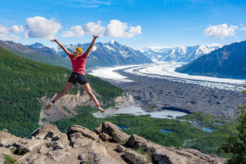 Young woman jumping into the air, view of impressive mountain landscape with Matanuska glacier and glaciated mountain peaks, Lion's Head, Chugach Mountains, Alaska, USA