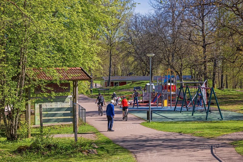 People cycling and walking by a playground with swings on a sunny spring day in a public park, Falköping, Sweden