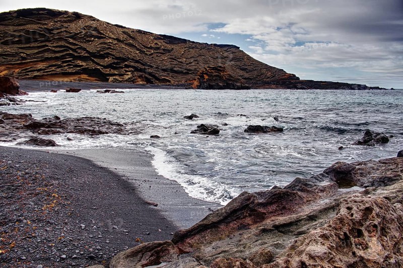 Secluded beach with black sand surrounded by wavy rock formations under a cloudy sky, Chargo Verde El Golfo Lanzarote