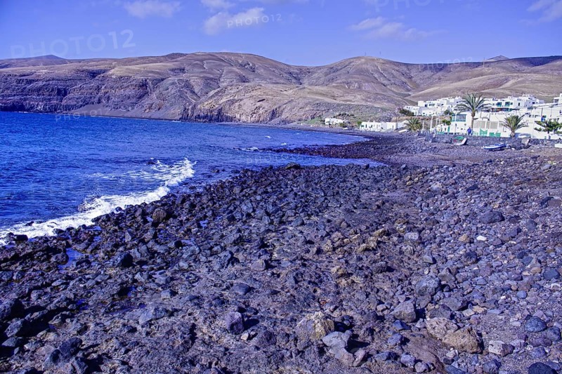 Rocky coastal landscape with blue waves and a village at the foot of the mountains under clear skies, Playa Quemada Yaiza Lanzarote