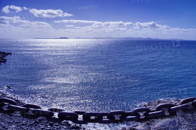 Chain on the edge of a cliff with a view of a calm, blue sea and distant island of Furteventura in the background, Playa Blanca Lanzarote