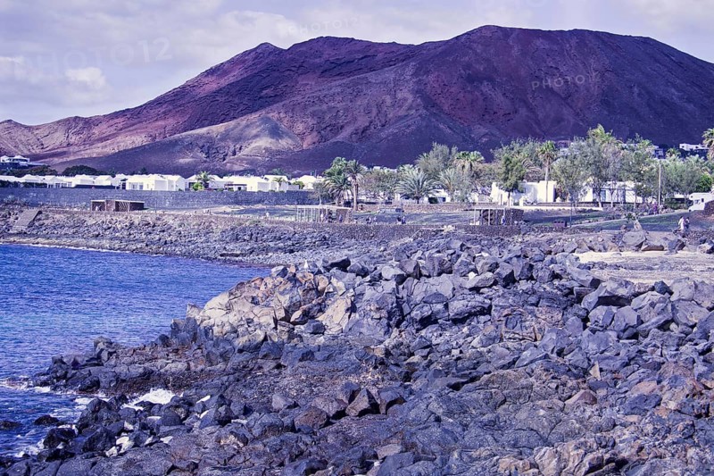 Rugged coastal area with rocks and views of reddish brown mountains under cloudy sky, Playa Blanca Lanzarote