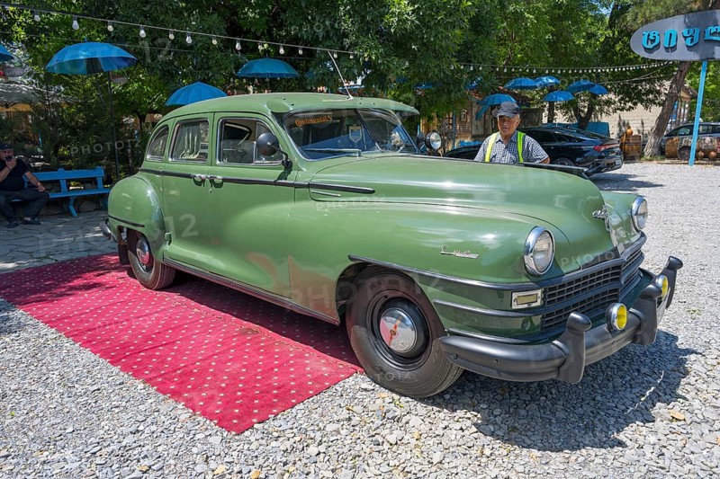 Green vintage car standing on outdoor red carpet surrounded by colorful umbrellas and gravel floor, vintage car, Chrysler Windsor limousine, Georgia