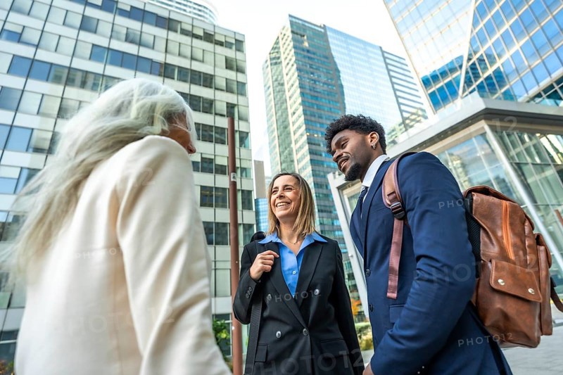 Diverse business professionals in smart casual attire chat and walk outside in a modern financial district, showcasing teamwork, collaboration and confident urban networking energy