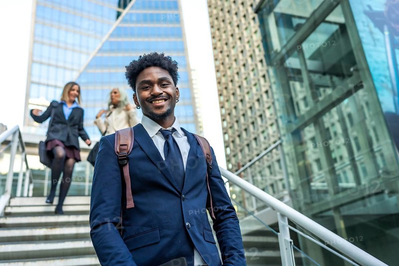 African american businessman smiling at camera and walking up stairs in a modern urban environment with female colleagues ascending steps in the background, conveying success and career growth