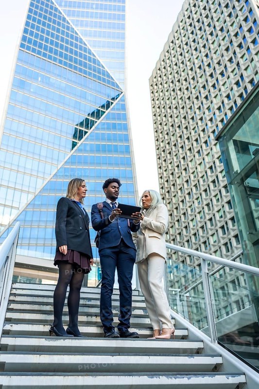 Diverse business team meeting on outdoor stairs in a downtown corporate district, collaborating over a tablet with skyscrapers behind, symbolizing teamwork and urban innovation