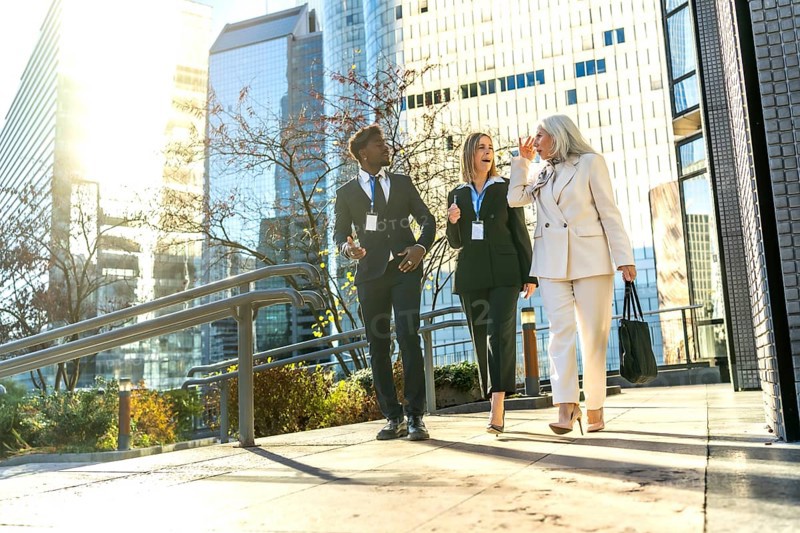 Three diverse business colleagues with name badges walking outdoors in a modern city discussing work, sharing ideas, and enjoying a conversation while commuting
