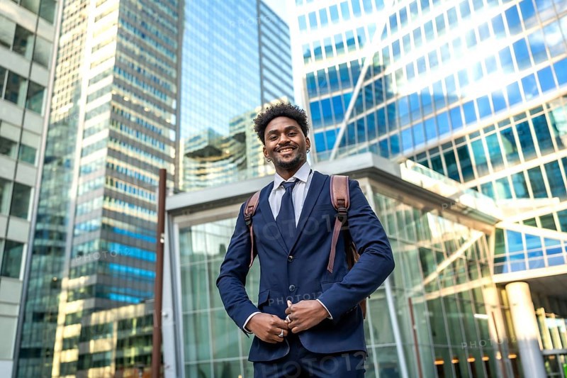 Young businessman standing confidently in a modern urban environment, carrying a backpack and smiling, representing success, ambition, and a dynamic professional lifestyle