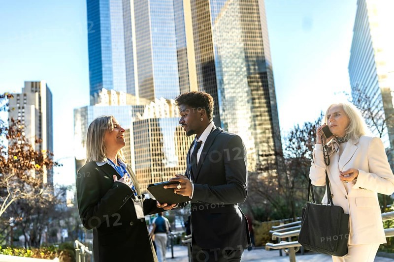 Diverse business professionals engaging in an outdoor meeting, sharing information on a tablet, and making a phone call, all set against a modern urban backdrop of towering skyscrapers