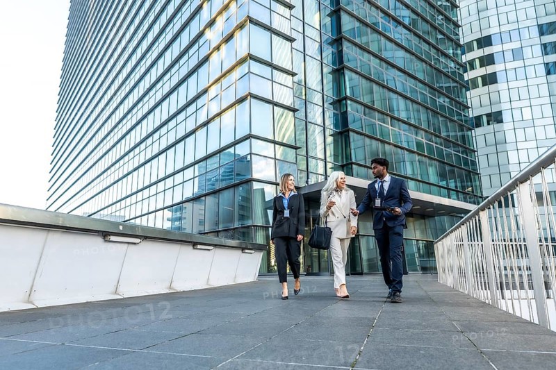 Business professionals of different ages and ethnicities are walking together, smiling, and having a discussion while leaving an urban corporate skyscraper