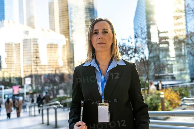 Professional businesswoman standing outdoors in a bustling city environment, wearing a suit and id badge while holding a device, representing corporate strength and urban success