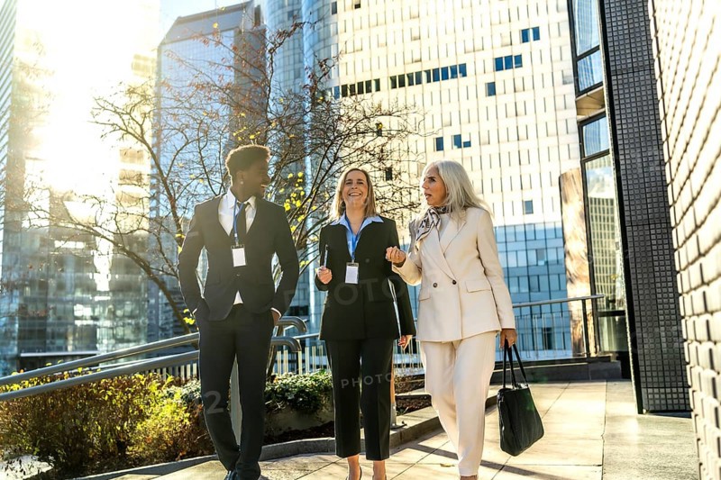 Diverse business professionals walking outdoors on a sunny day, engaging in conversation while heading to work in a modern corporate urban environment with skyscrapers