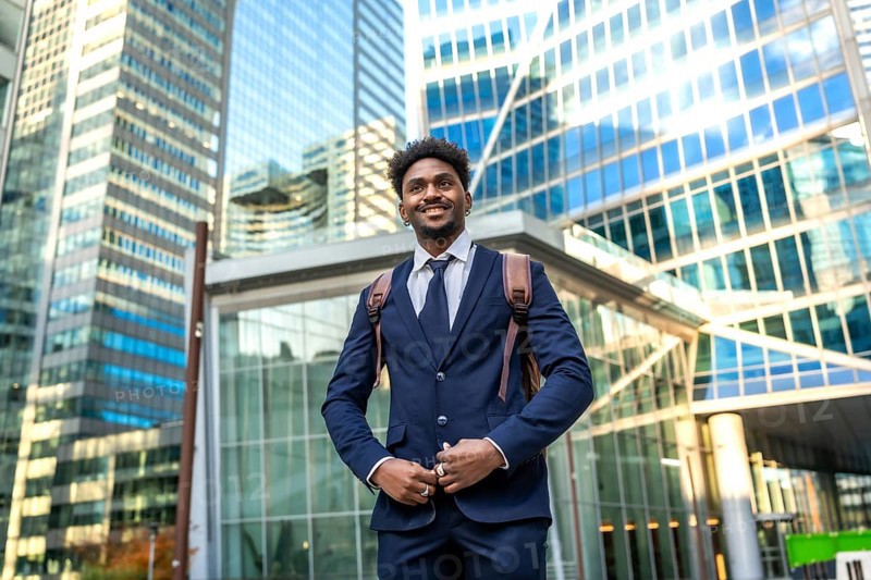 Young african american businessman smiling, looking ahead while standing confidently with a backpack in front of gleaming corporate buildings, representing success and urban professionalism