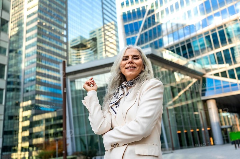 Senior businesswoman with gray hair and a formal blazer smiling confidently, standing outdoors against a backdrop of modern glass skyscrapers in a bustling city