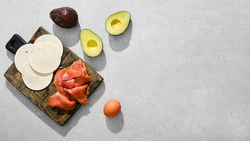 Smoked salmon, slices of goat cheese, halved avocados, and an egg on a wooden cutting board. Healthy food ingredients