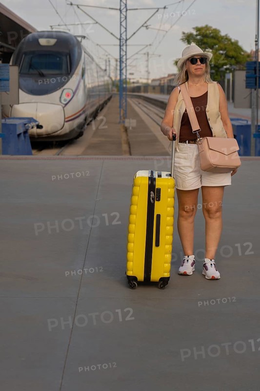 Adult female traveler standing on a railway platform with a yellow suitcase, looking around while waiting for her train in a modern city station