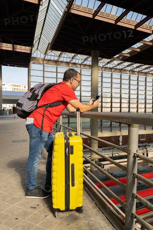 Adult man leaning on a railing at a modern city train station, checking his smartphone while standing next to a yellow suitcase during an urban travel moment