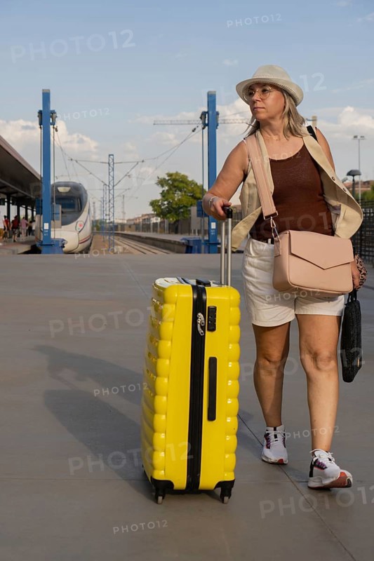 Adult woman traveling alone at a city train station, pulling a yellow suitcase while walking along the platform during a relaxed daytime journey