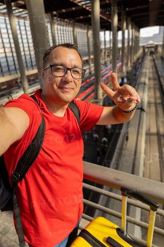 Middle aged man wearing reading glasses smiles calmly while taking a selfie and making a peace sign at a modern train station. Relaxed travel and urban lifestyle concept