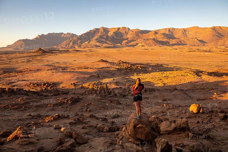 Young woman standing in desert landscape with Brandberg in morning light, at sunrise, Erongo, Damaraland, Namibia
