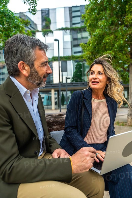 Two professional business colleagues, a man and a woman, are sitting on a bench in a city environment, collaborating and having an informal discussion while using a laptop