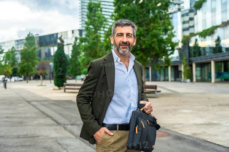 Confident mature businessman carrying a briefcase and smiling at the camera, standing outdoors with one hand in his pocket against a modern urban background, representing success and professionalism