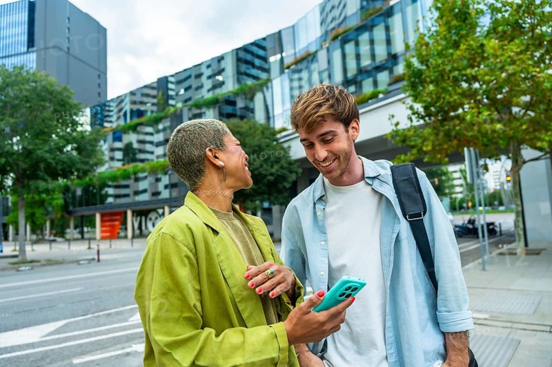 Two diverse colleagues enjoying a casual conversation and sharing a smartphone screen on an urban street, featuring modern city architecture and green spaces