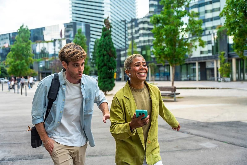 Diverse business colleagues walking on a paved pathway outdoors in a modern city district, sharing a joyful moment and communicating while carrying a shoulder bag and smartphone