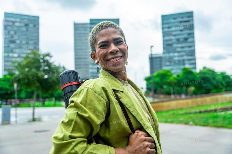 Urban architect smiling confidently with drawing tube on shoulder strap, standing in front of modern downtown skyscrapers, conveying creative success, ambition and forward thinking design