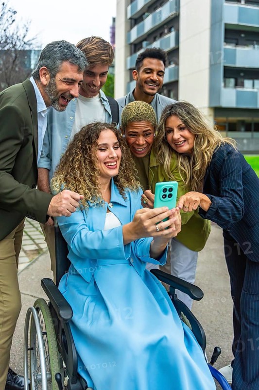 Diverse group of professional colleagues, including a woman in a wheelchair, smiling and interacting around a mobile phone, embracing inclusion and technology in an urban setting