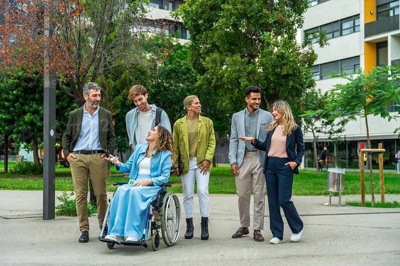 Diverse group of business people, including a woman in a wheelchair, engaging in a lively outdoor discussion surrounded by greenery and modern city buildings, promoting inclusion and teamwork