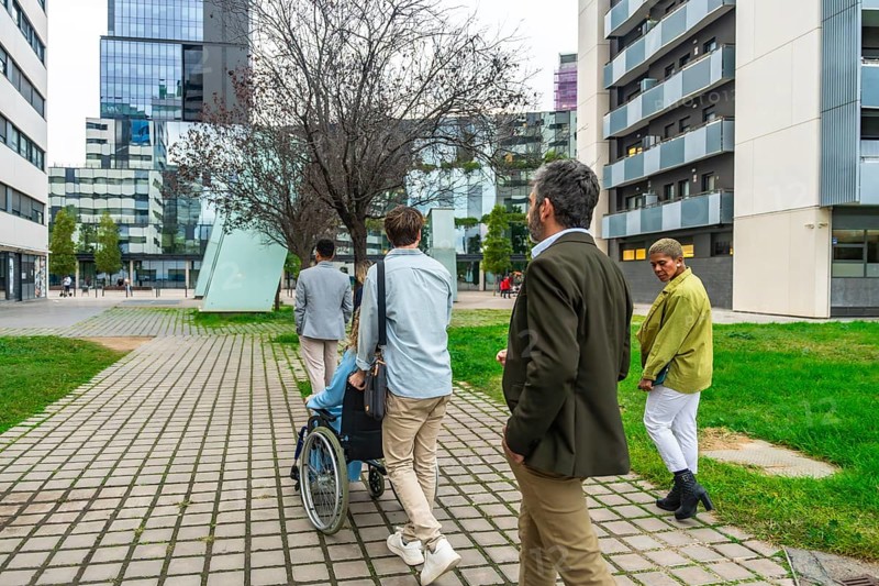 Diverse group of colleagues walking on a paved pathway through a modern city district, one person pushing a colleague in a wheelchair, symbolizing inclusivity and teamwork
