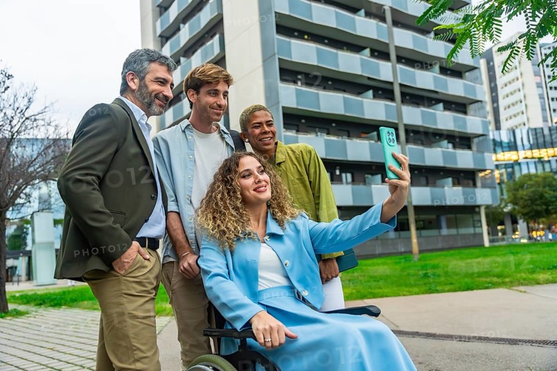 Diverse group of smiling business colleagues, including a woman in a wheelchair, capturing a selfie together with a smartphone outdoors in a modern city setting