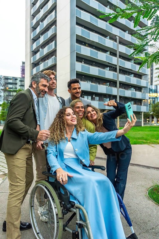 Group of multi ethnic colleagues and friends, including a woman in a wheelchair, smiling and taking a cheerful outdoor selfie in a modern urban setting, showing inclusion and teamwork