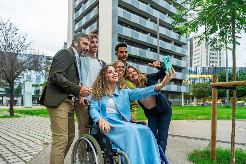 Diverse group of business colleagues, including a woman in a wheelchair, smiling and taking a selfie with a smartphone outdoors in a city setting, showcasing teamwork and inclusion