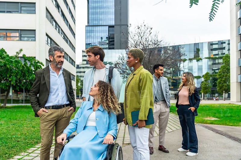 Group of diverse business professionals, including a woman using a wheelchair, collaborating and communicating in a modern urban park setting between office buildings