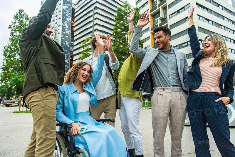 Diverse business colleagues including a woman in a wheelchair cheering and doing high fives outdoors in a modern city, celebrating teamwork, inclusion, accessibility, and achievement