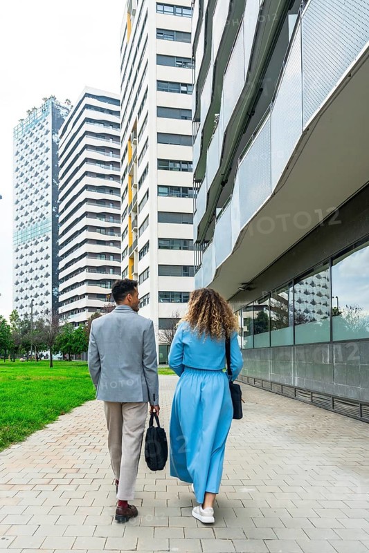 Two business professionals walking together along a paved pathway in an urban area, passing tall contemporary buildings, representing teamwork, partnership, and city life