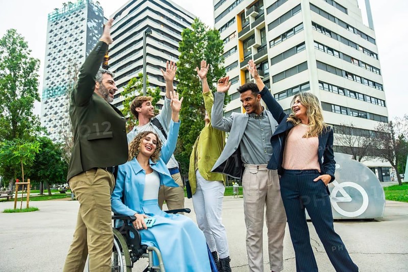 Mixed age businesspeople with a woman in a wheelchair raising their hands in celebration, showing unity, teamwork, and inclusion in a modern city setting with buildings and green space
