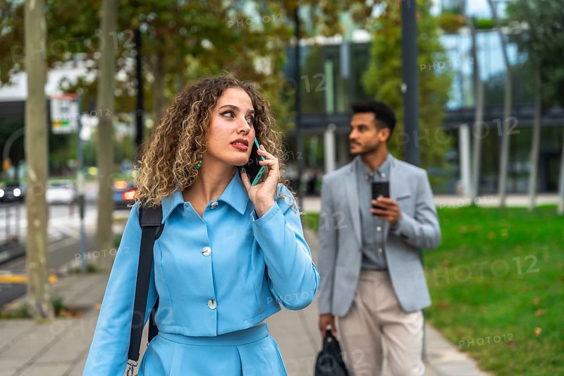 Confident businesswoman talking on her smartphone while walking on a city sidewalk with a male colleague carrying a laptop bag in the background, representing modern urban professionals