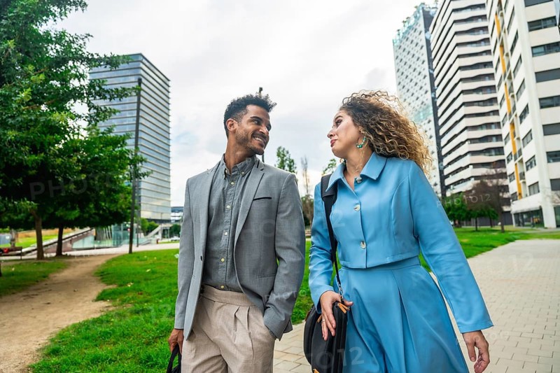 Two diverse business professionals walking along a path in a contemporary urban park with modern buildings, engaging in conversation while collaborating outdoors