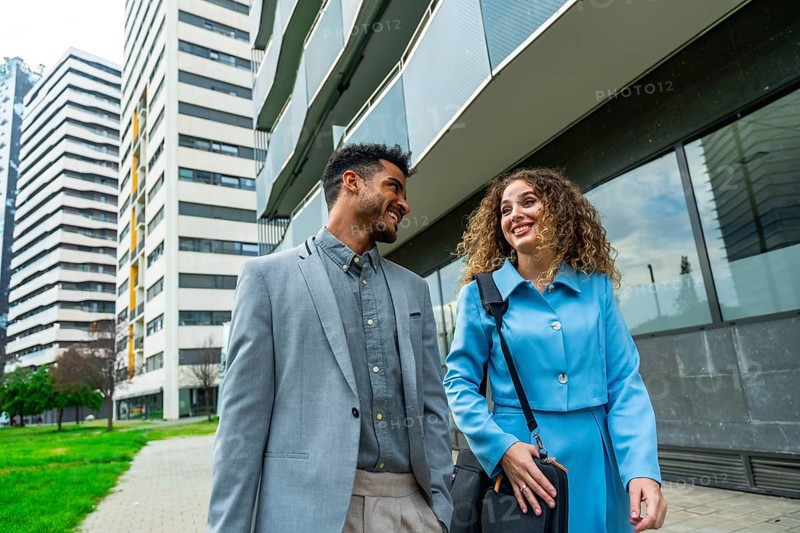 Two diverse business professionals walking outside a modern office building, looking at each other and smiling, embodying teamwork and professional camaraderie in an urban environment