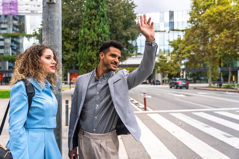 Young businessman hails a taxi on a busy downtown sidewalk while his female colleague stands beside him, both in smart casual attire amid modern city architecture and traffic