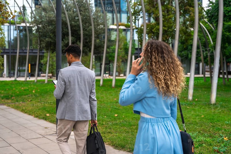 Two diverse business colleagues, a man texting on his smartphone and a woman conversing on hers, walking outdoors in a urban park setting during a workday