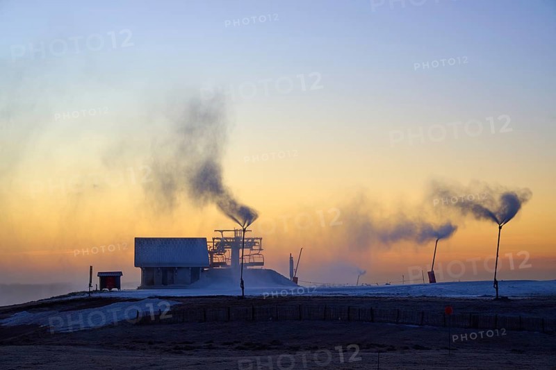 A factory with snow cannons emits smoke under a blue and yellow evening sky, Winter, Route de Cretes, Hohneck, La Bresse, Vosges, France