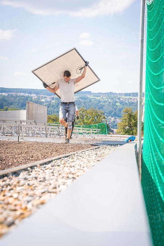 Man carrying a solar panel across a building site with a view and green fence, new park and ride parking garage Calw Heumaden on the new Hermann Hessebahn, Calw, Germany