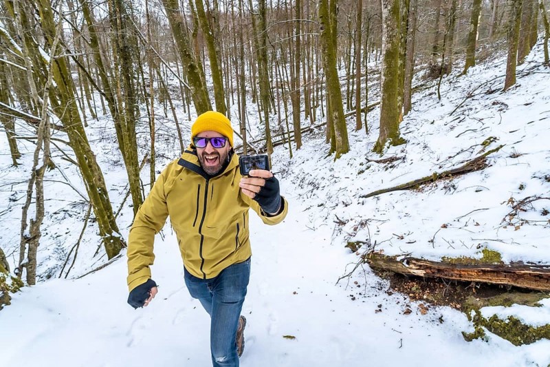 Happy man in a yellow beanie and green jacket smiling at his action camera while vlogging on a snowy forest hiking trail, capturing winter adventure and outdoor fun